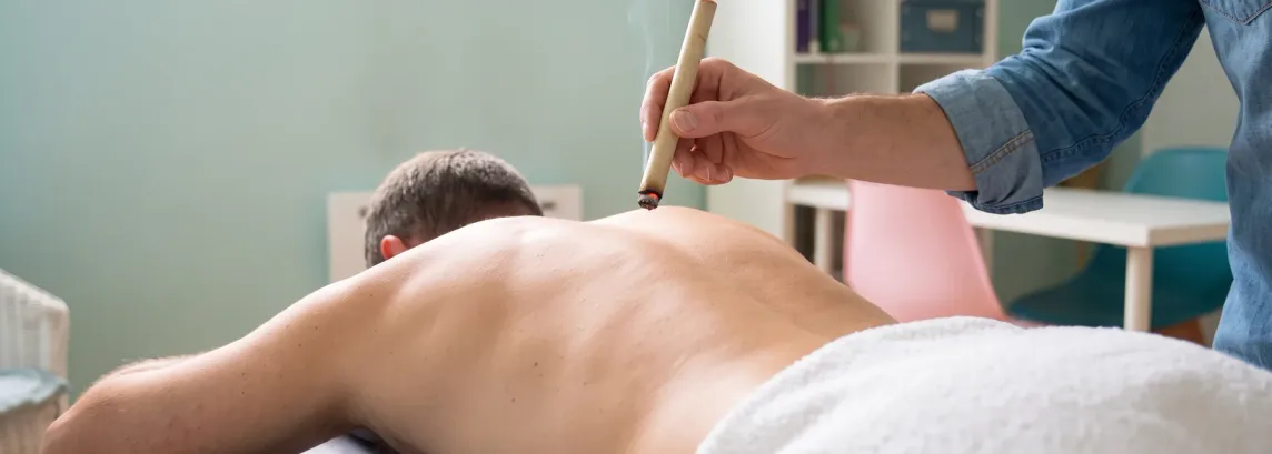 Une personne allongée sur le ventre sur la table de soin, une serviette blanche sur le bas du dos. La main du praticien, manche en jean retroussée, tient un bâton de moxa allumé à quelques centimètres au-dessus de ses omoplates.